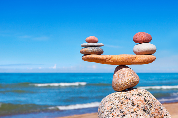 Several stones balanced on top of each other with a beach and wavy sea in the background, as well as blue sky.